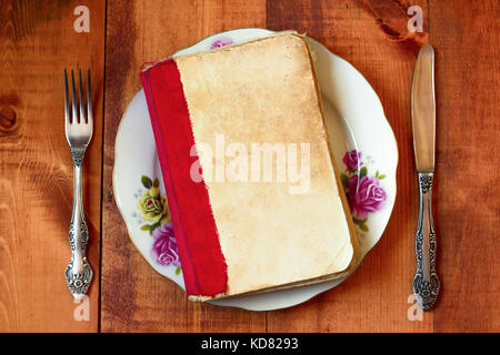 Book on a plate with fork and knife on wooden table. Concept image of a book as main course meaning 'feeding with culture' Stock Photo