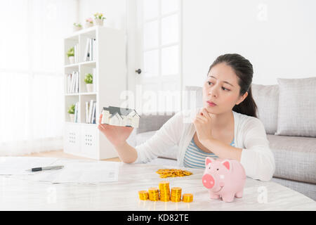 beautiful pretty girl lady holding little house model looking at thinking how to saving money to buying new apartment sitting on living room with pigg Stock Photo