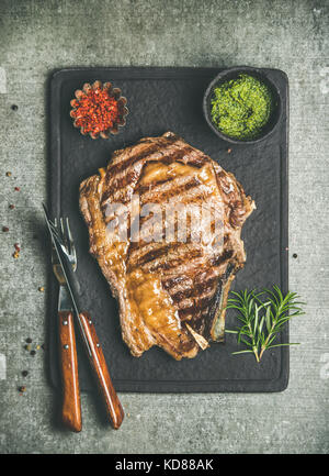 Flat-lay of grilled hot rib-eye beef meat steak on bone with chimichurri green sauce and hot red chili pepper flakes on black slate serving board over Stock Photo
