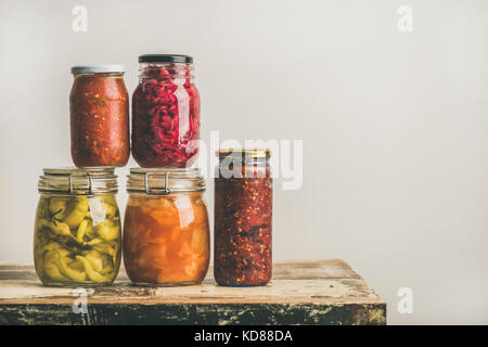 Autumn seasonal pickled or fermented colorful vegetables in glass jars placed in stack over vintage kitchen drawer, white wall background, copy space. Stock Photo