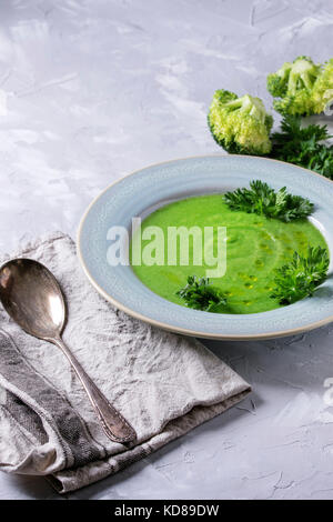 Vegetarian vegan broccoli cream soup served in blue plate with fresh parsley, broccoli, spoon, textile napkin over gray concrete background. Healthy e Stock Photo