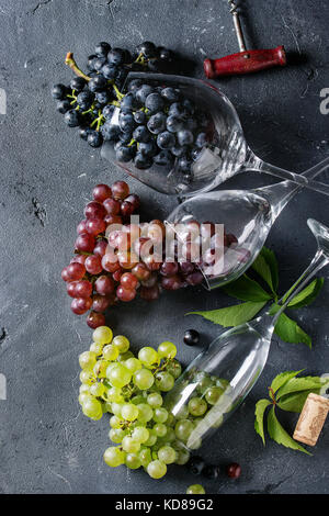 Variety of three type fresh ripe grapes dark blue, red and green in different lying wine glasses with old corkscrew over black texture background. Top Stock Photo