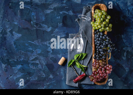 Variety of three type fresh ripe grapes dark blue, red and green in wooden bowl with empty laying wine glass, old corkscrew and green leaves over blue