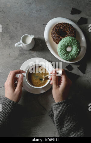 Plate of colorful glazed donuts with chopped chocolate, cup of black coffee, jug of milk over gray texture table. Female hands. Flat lay with space. R Stock Photo