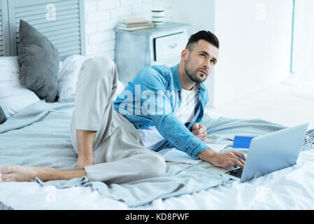 Pensive freelancer lying in bed while working from home Stock Photo