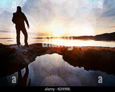 Lens defect.  Tall backpacker watch clear sunny spring daybreak over sea. Hiker with backpack stand on rocky shore and his figure is mirrored in water Stock Photo