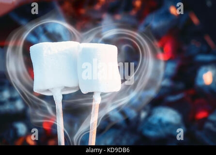 Two white sweet marshmallows roasting over fire flames. Smoke in form of heart. Marshmallow on skewers roasted on charcoals Stock Photo