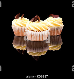Three muffin cupcake with yellow cream and pieces of chocolate in a paper cup. On a black glossy background with real reflection. Isolated on black Stock Photo