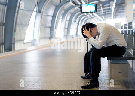 Train delay or problem in the train station, tired desperate Aisan passenger waiting in the terminal with suitcase. Stock Photo