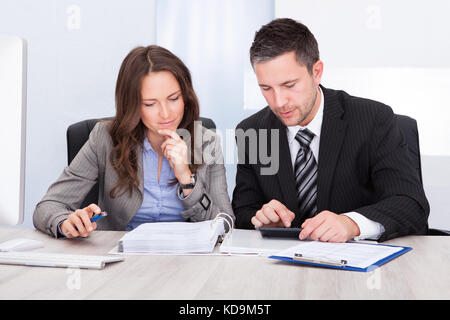 Two Young Businesspeople Calculating Bills At Desk In Office Stock Photo