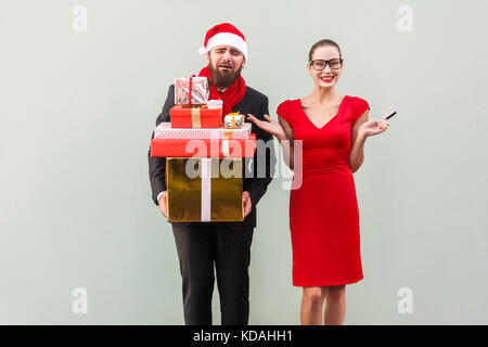 Unhappy bearded well dressed man holding many gift box and crying, women in black glasses and red dress looking at camera, toothy smiling and hands on Stock Photo