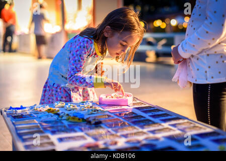 Little girl is picking jewellery at traditional Mediterranean market stand. Child is choosing handmade rings, necklaces and beads in the evening. Stock Photo