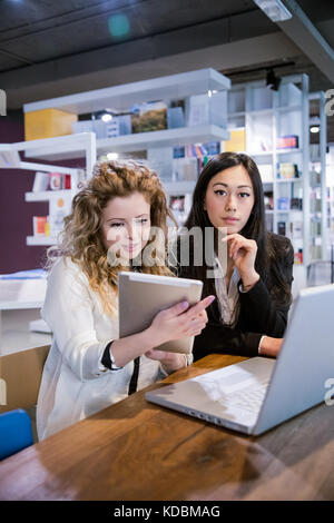 two female coworkers discussing with each other using tablet on table in design studio Stock Photo