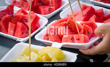 Fresh watermelon and pineapple slice in white foam plate container on black wooden table. Serving by one hand. Stock Photo