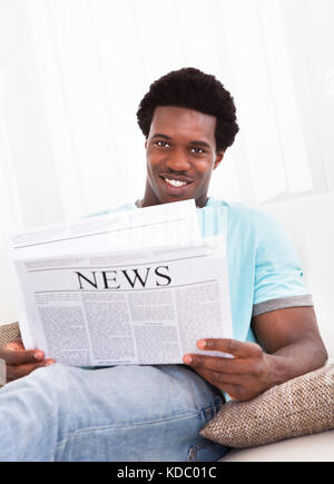 Portrait Of Casual Young African Man Reading Newspaper Sitting On Couch Stock Photo