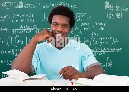 Young African Man Studying With A Lot Of Books Sitting In Front Of Blackboard Stock Photo