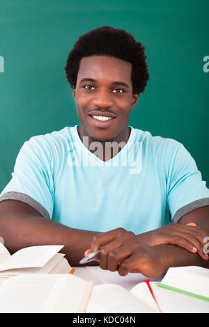 Young African Man Studying With A Lot Of Books Sitting In Front Of Blackboard Stock Photo