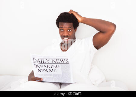 Portrait Of Worried Young African Man Holding Newspaper Sitting On Bed Stock Photo