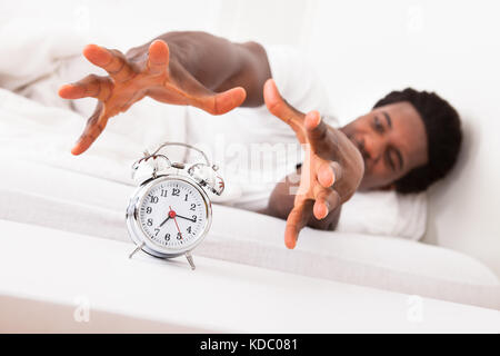 Frustrated Man Trying To Catch Is Alarm Clock While Relaxing On His Bed Stock Photo