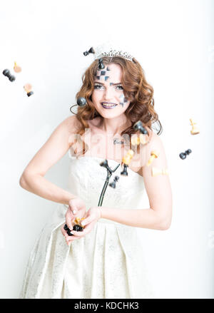 Young attractive woman with face painted to chess light and dark squares dressed in white dress and crown throwing up chess figures into air Stock Photo
