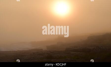 Mist and clouds covering the air, very low visibility, during sunset in Hermanus, South Africa, part of Garden Route. Golden sunset by the coast waves Stock Photo