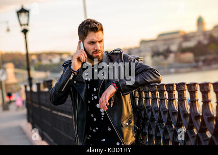 A handsome young hipster man leaning to the fences in the sunset while talking on his smartphone Stock Photo