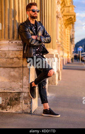 A handsome young hipster man leaning to the pillar of a house on a sunny street and relaxing with his sunglasses on Stock Photo