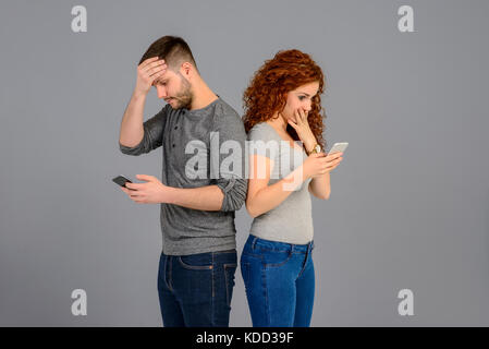 A beautiful young couple standing back to back while holding their smartphones and feeling surprised Stock Photo