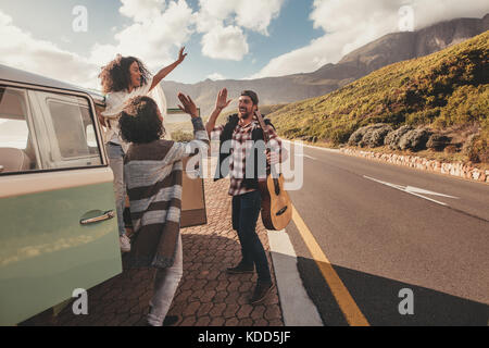Group of man and women on road trip standing by the van and giving high five. Cheerful friends enjoying themselves on a vacation. Stock Photo