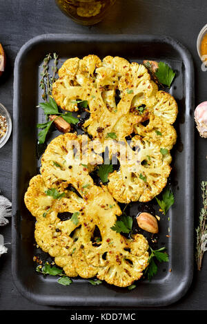 Roasted cauliflower steaks with herbs and spices on baking sheet over black stone background, top view Stock Photo