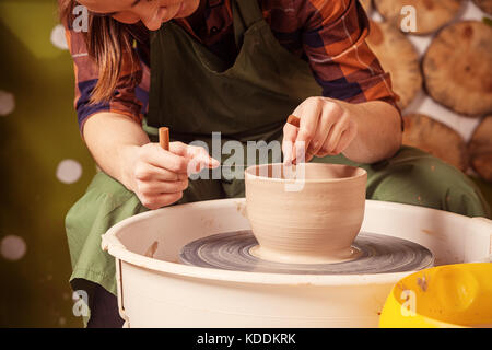 A potter in a plaid shirt and green apron beautifully sculpts a deep bowl of brown clay and cuts off excess clay on a potter's wheel in a beautiful wo Stock Photo