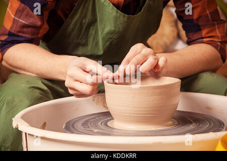 Close-up A woman potter in a plaid shirt and green apron beautifully sculpts a deep bowl of brown clay and cuts off excess clay on a potter's wheel in Stock Photo