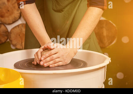 A young woman in a plaid shirt and jeans sculpts a clay bowl on a potter's wheel in a bright beautiful workshop decorated with wooden circles Stock Photo