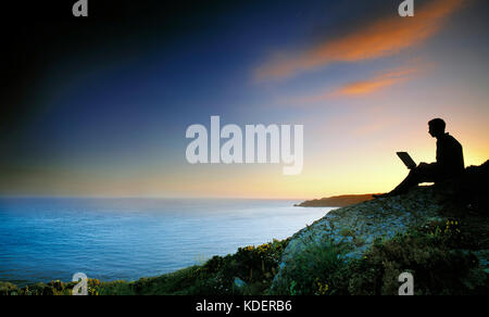 Man working on laptop computer whilst sitting on coastal clifftop at sunset. Stock Photo