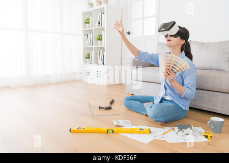 smiling young woman wearing VR technology goggles viewing new house design sketch drawing and touching simulation screen to choose color. Stock Photo