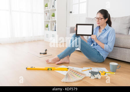 pleasantly attractive female college design department student doing homework in living room wooden floor and holding mobile pad computer showing blan Stock Photo