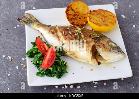 Grilled dorado with grilled lemon, spinach and tomato on a white plate on a grey abstract background. Healthy eating concept. Mediterranean lifestyle Stock Photo