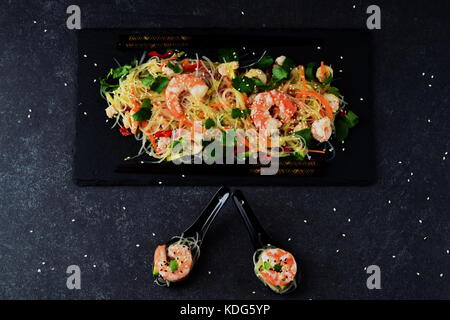 Noodles with prawns and vegetables on a black stone plate and in traditional soup spoons on a grey abstract background. Healthy food concept Stock Photo