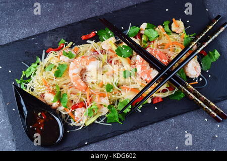 Noodles with prawns and vegetables on a black stone plate and in traditional soup spoons on a grey abstract background. Healthy food concept Stock Photo