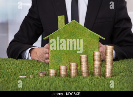 Businessman Holding Eco Friendly House In Front Of Stack Of Coins Over Grass Stock Photo