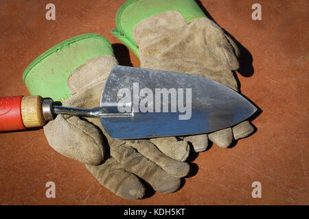 A pair of dirt-stained leather work gloves lies on a brown table top with a silver garden trowel  with red wooden handle laid on top of them. Stock Photo