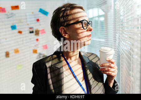 Mature officer with glass of coffee looking through venetian blinds on window Stock Photo