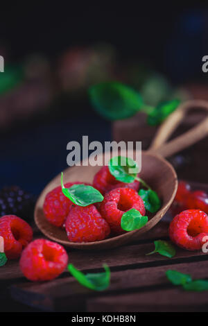 Summer still life with berries in a wide wooden spoon on a dark background. Raspberry with green mint leaves. Stock Photo