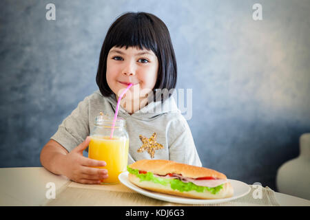 Cute black hair little girl drinking fresh  orange juice and eating sandwich Stock Photo