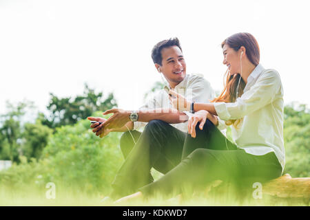 Young Asian lovely couple or college student sit listening to song music on smartphone together in park. Leisure activity, dating relationship concept Stock Photo