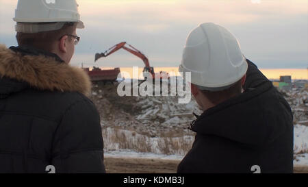 Back view of architects inspecting construction site. Two engineers in hard hats on construction site excavator in the background. Engineers examining documents at construction site against excavator. Stock Photo