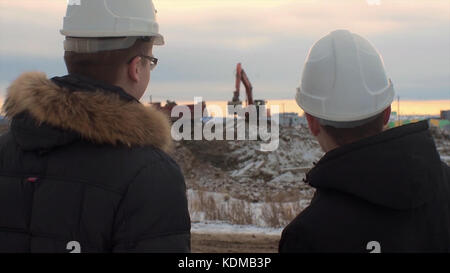 Back view of architects inspecting construction site. Two engineers in hard hats on construction site excavator in the background. Engineers examining documents at construction site against excavator. Stock Photo