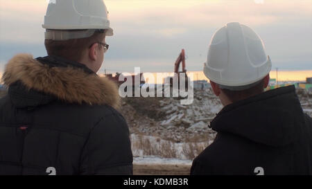 Back view of architects inspecting construction site. Two engineers in hard hats on construction site excavator in the background. Engineers examining documents at construction site against excavator. Stock Photo