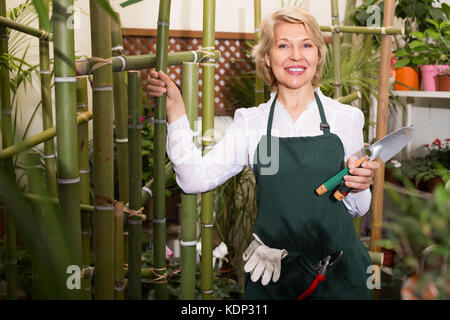Joyful elderly female florist with tools in hands among green plants in floral shop Stock Photo