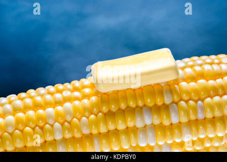 Closeup of a steaming organic corn cob with a piece of melting butter on top. Blue background. Stock Photo